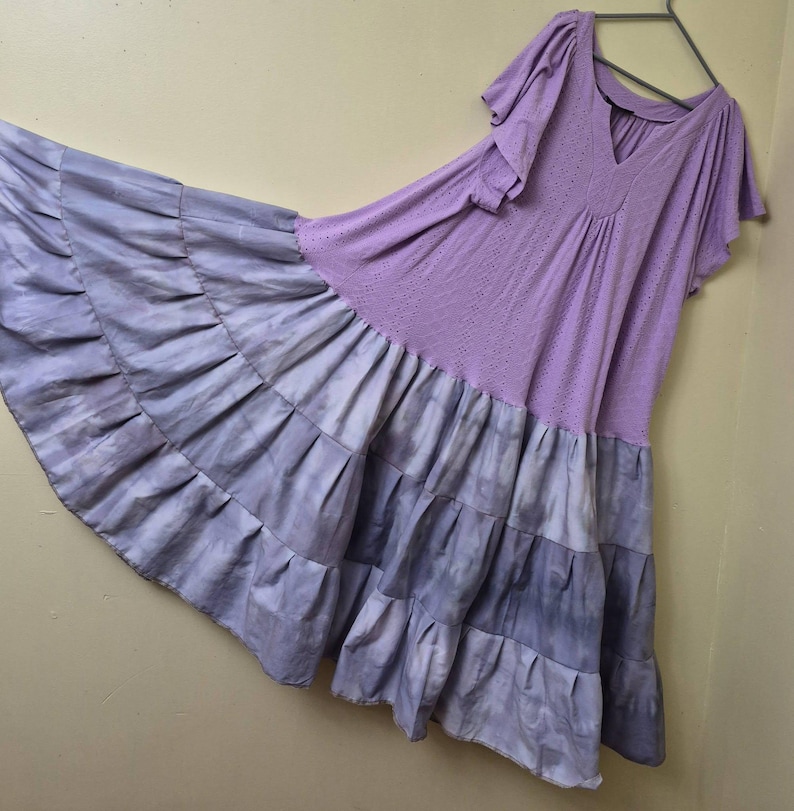 May include: A flowing, lavender dress with short sleeves and a V-neck. The bodice has a perforated design, and the skirt features layered ruffles in shades of lavender and gray. The dress is hanging on a metal hanger.