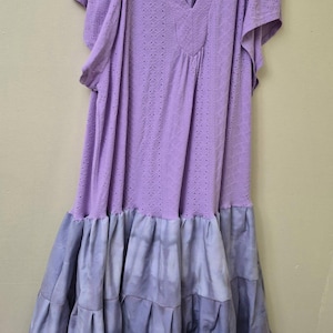 May include: A lavender dress with short sleeves and a V-neckline. The top portion has a perforated design, while the skirt features a tiered, tie-dye effect in shades of blue and purple. The dress is hanging on a gray hanger.