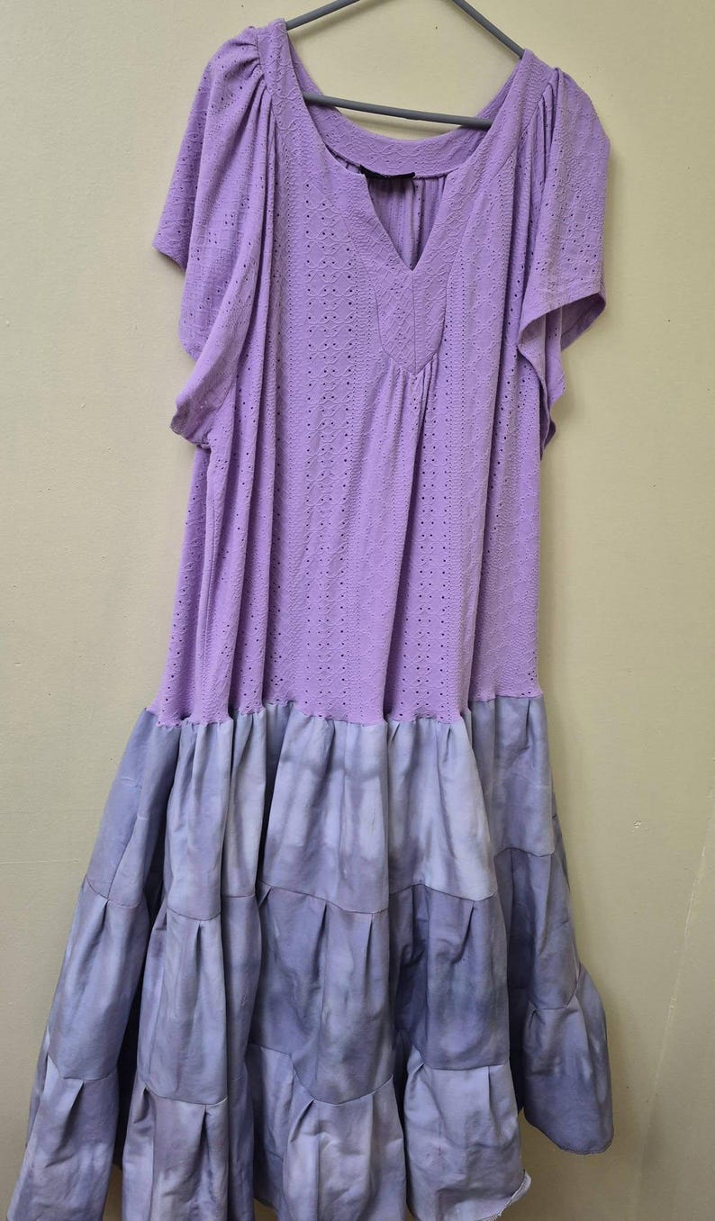 May include: A lavender dress with short sleeves and a V-neckline. The top portion has a perforated design, while the skirt features a tiered, tie-dye effect in shades of blue and purple. The dress is hanging on a gray hanger.