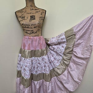 May include: A long, tiered skirt featuring alternating bands of solid dusty pink, metallic gold, and a white fabric with a small brown floral print. The hem is finished with a wide panel of light pink fabric featuring a white damask-style pattern.