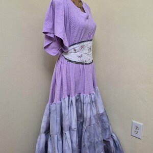 May include: A lilac-colored dress with a tiered skirt and a floral patterned belt. The dress has short sleeves and a scoop neckline. The skirt transitions from lilac to a gray-blue hue. The dress is displayed on a mannequin.