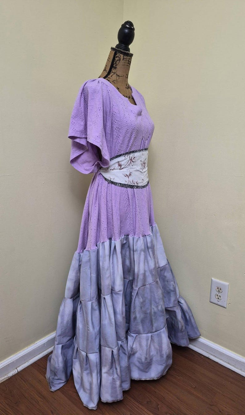 May include: A lilac-colored dress with a tiered skirt and a floral patterned belt. The dress has short sleeves and a scoop neckline. The skirt transitions from lilac to a gray-blue hue. The dress is displayed on a mannequin.