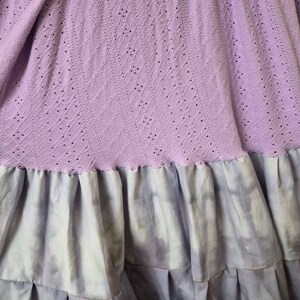 May include: A lilac-colored dress with a detailed eyelet bodice and long sleeves. The skirt features a tie-dye effect in shades of lavender and gray, with multiple pleated ruffles. The dress is made of soft fabric.