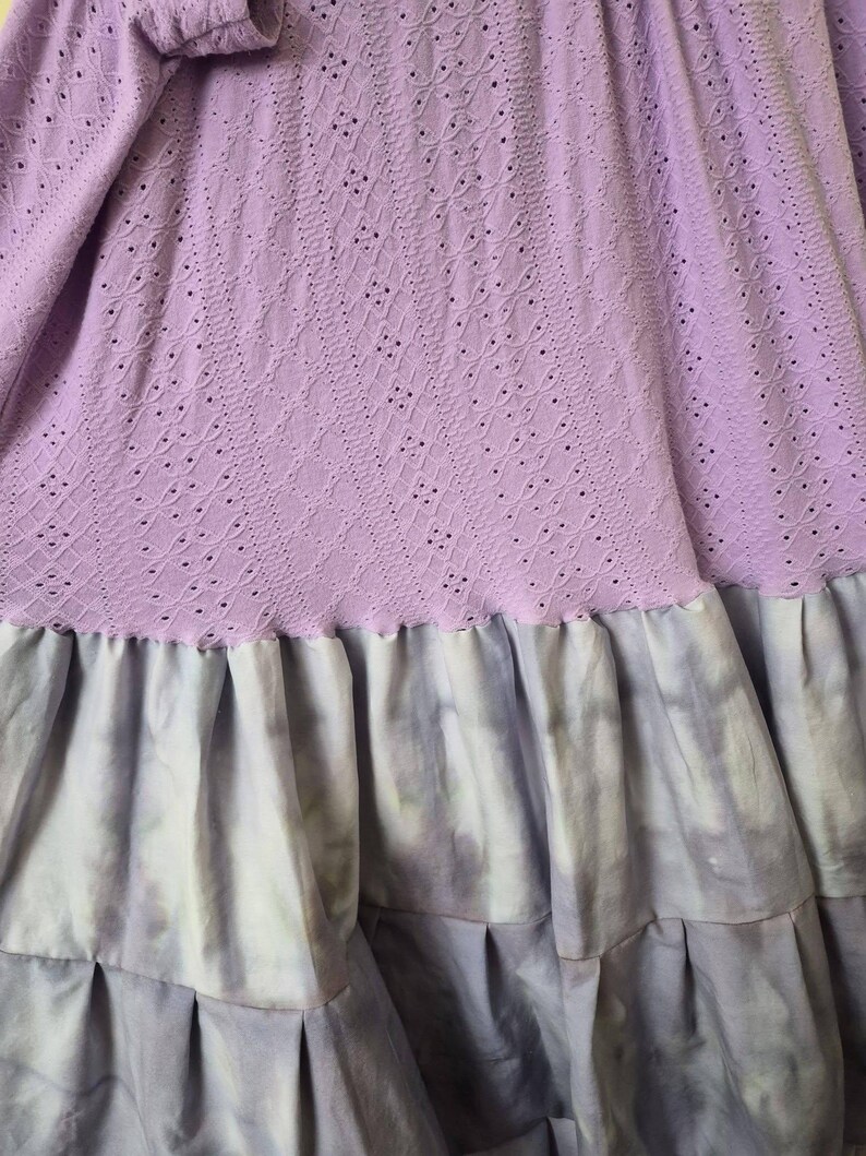 May include: A lilac-colored dress with a detailed eyelet bodice and long sleeves. The skirt features a tie-dye effect in shades of lavender and gray, with multiple pleated ruffles. The dress is made of soft fabric.