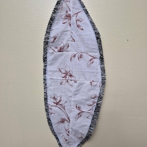 May include: A leaf-shaped fabric item with a floral pattern in maroon on a white background. The edges are trimmed with a black, fringed material. A maroon fabric loop extends from the bottom.