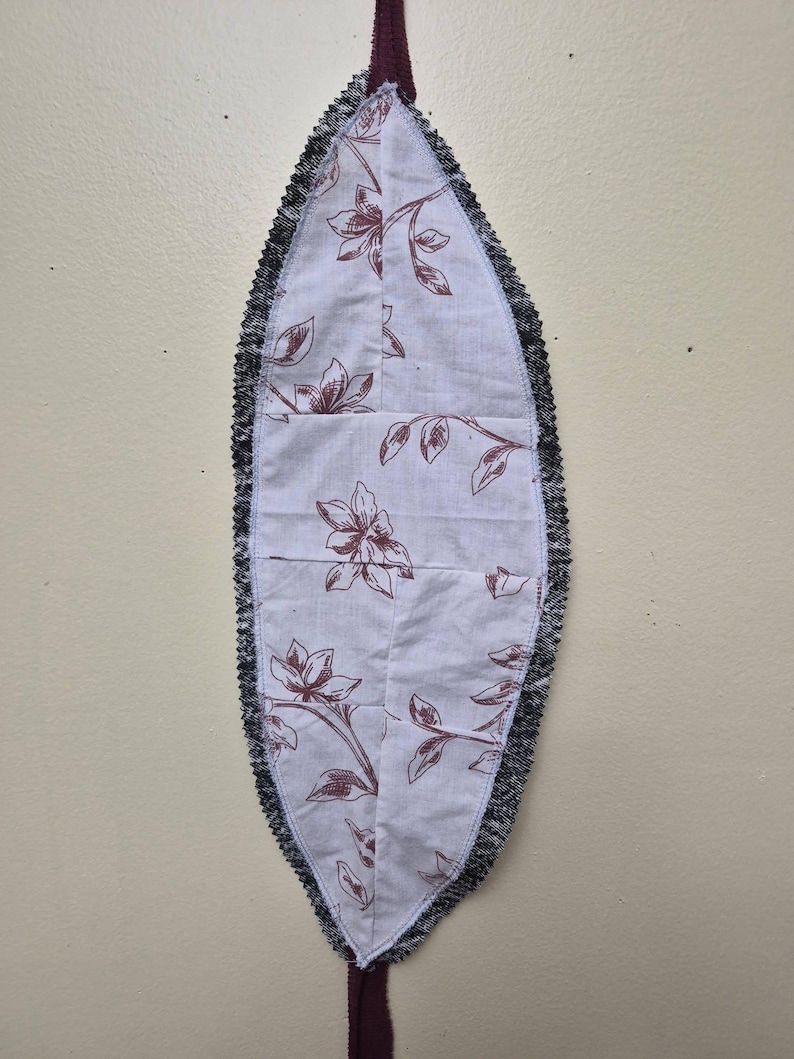 May include: A leaf-shaped fabric item with a floral pattern in maroon on a white background. The edges are trimmed with a black, fringed material. A maroon fabric loop extends from the bottom.