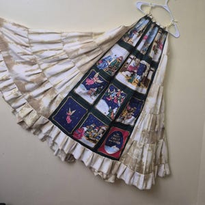 May include: A tiered skirt featuring a central panel with rectangular illustrations of nativity scenes and angels on a dark green background. The text "THE NIGHT CHRIST WAS BORN" is printed on one panel. The sides are beige with a mottled pattern.