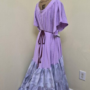 May include: A long, flowing dress in shades of lavender and gray, displayed on a mannequin. The bodice is a light purple with a textured, eyelet design, and the skirt transitions to a tiered, tie-dye effect in gray. A dark brown belt cinches the waist.