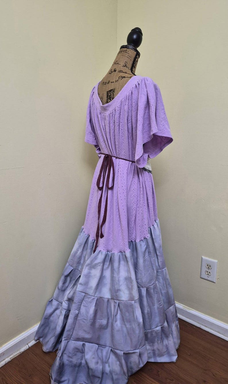 May include: A long, flowing dress in shades of lavender and gray, displayed on a mannequin. The bodice is a light purple with a textured, eyelet design, and the skirt transitions to a tiered, tie-dye effect in gray. A dark brown belt cinches the waist.