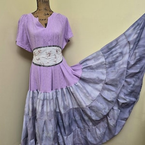May include: A lilac-colored dress with a V-neck and short sleeves. The dress features a floral patterned belt and a tiered, tie-dye skirt in shades of purple and gray. The dress is displayed on a mannequin.