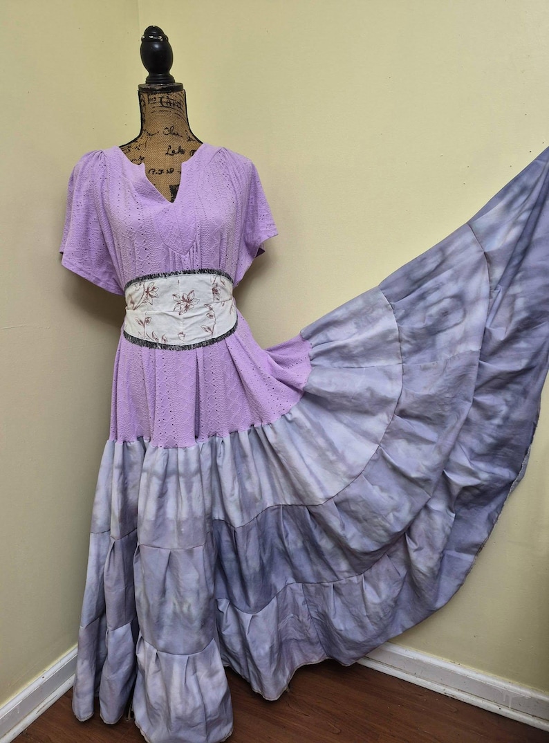 May include: A lilac-colored dress with a V-neck and short sleeves. The dress features a floral patterned belt and a tiered, tie-dye skirt in shades of purple and gray. The dress is displayed on a mannequin.