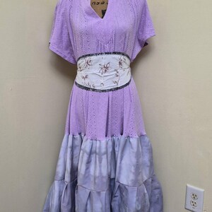May include: A lilac-colored dress with a V-neck and short sleeves. The dress features a floral patterned belt and a tiered, tie-dye effect skirt. The dress is displayed on a mannequin.