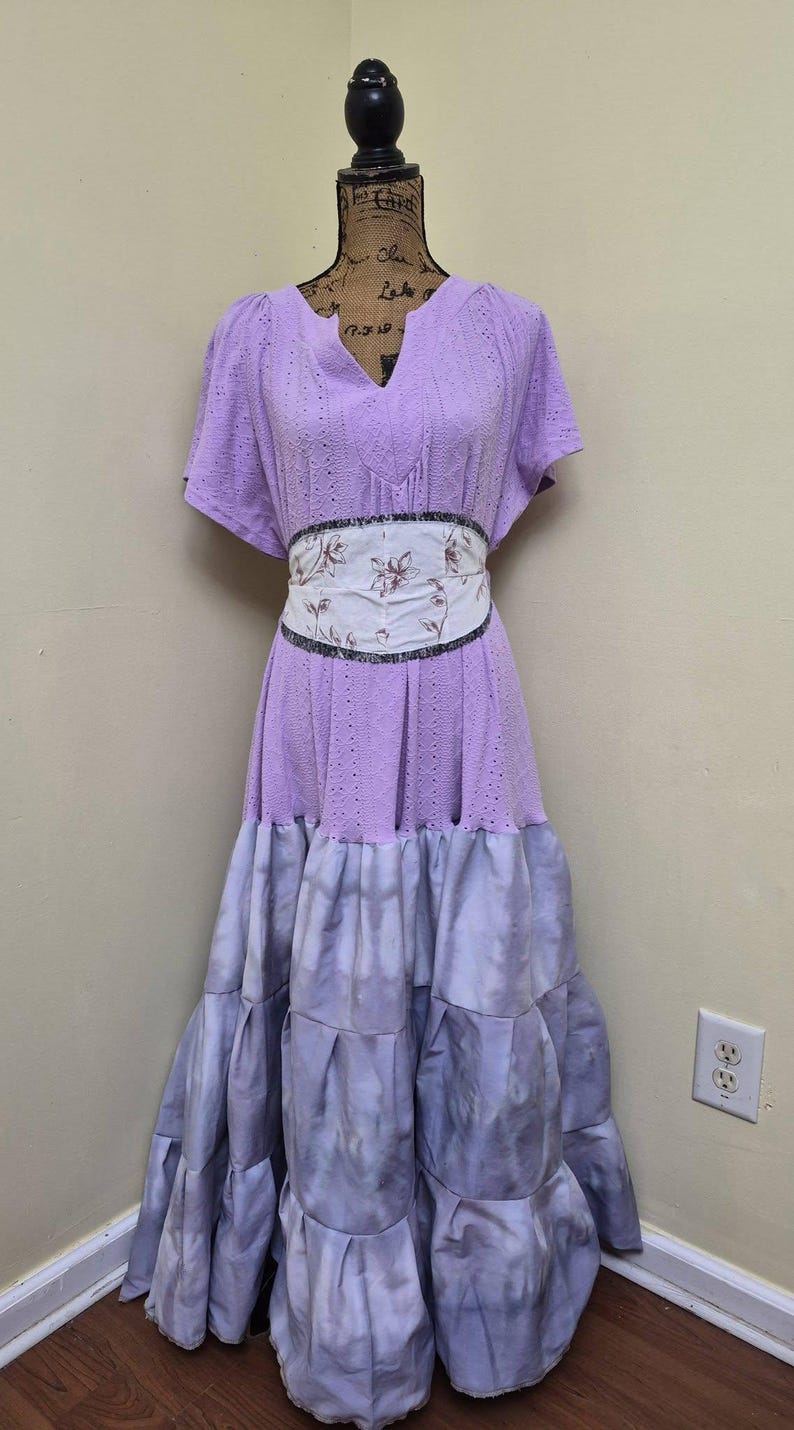 May include: A lilac-colored dress with a V-neck and short sleeves. The dress features a floral patterned belt and a tiered, tie-dye effect skirt. The dress is displayed on a mannequin.