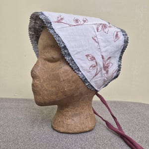 May include: A white bonnet with a floral pattern in a muted red hue. The bonnet has a dark gray and black trim and is tied with long, burgundy-colored ribbons. The bonnet is displayed on a mannequin head.