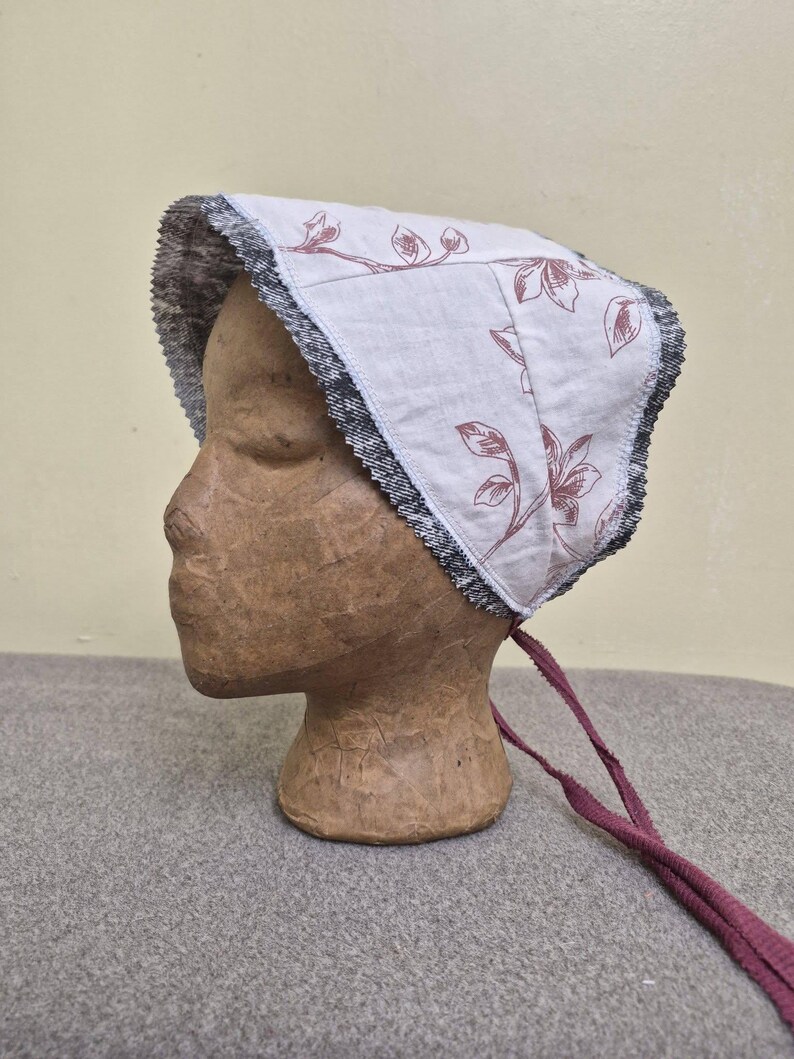 May include: A white bonnet with a floral pattern in a muted red hue. The bonnet has a dark gray and black trim and is tied with long, burgundy-colored ribbons. The bonnet is displayed on a mannequin head.