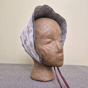 May include: A vintage-style bonnet with a floral pattern on a cream background. The bonnet has a dark brown textured brim and is trimmed with a black and white border. Burgundy ribbons hang from the bonnet. The bonnet is displayed on a mannequin head.