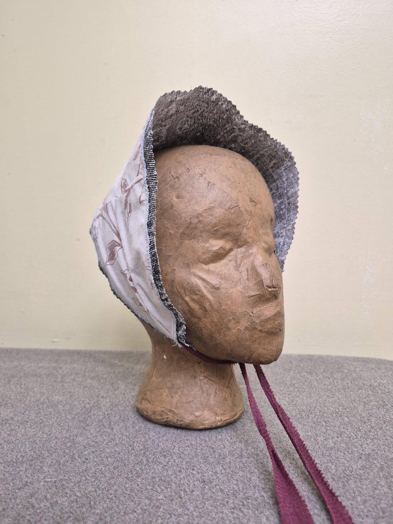 May include: A vintage-style bonnet with a floral pattern on a cream background. The bonnet has a dark brown textured brim and is trimmed with a black and white border. Burgundy ribbons hang from the bonnet. The bonnet is displayed on a mannequin head.