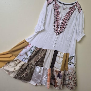May include: A white knee-length dress featuring a V-neckline with dark pink and black embroidery. The bodice has a button-down front and short sleeves. The skirt is constructed from a patchwork of various fabrics in mustard yellow, brown, and floral prints.