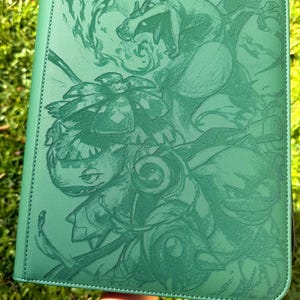 Könnte beinhalten: Ein rechteckiger grüner Ordner mit einer detaillierten, einfarbigen Gravur von drei Kreaturen, darunter ein feuerspeiender Drache, ein pflanzenartiges Wesen mit einer großen Blüte und eine schildkrötenartige Kreatur mit Kanonen.