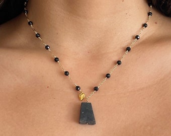 May include: A gold chain necklace with black beads and a dark gray, rectangular pendant. A small gold heart charm is also attached. The necklace is worn around the neck.
