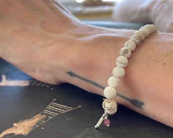 May include: A white and brown beaded bracelet with a small silver charm and a pink gemstone. The bracelet is on a person's wrist.