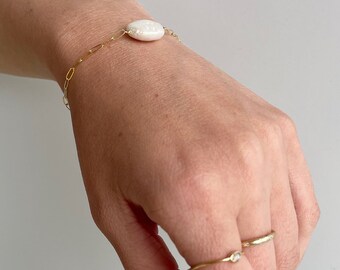 May include: A gold chain bracelet with a single white pearl charm. The bracelet is worn on a person's wrist. The person is also wearing two gold rings with small white stones on their finger.