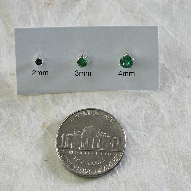 May include: Three green gemstone earrings on a white card with measurements in millimeters: 2mm, 3mm, and 4mm. A US quarter is shown for size comparison.