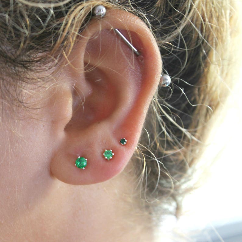 May include: Close-up of a person's ear with multiple piercings. The ear is pierced with a silver industrial barbell and three small studs with green gemstones.