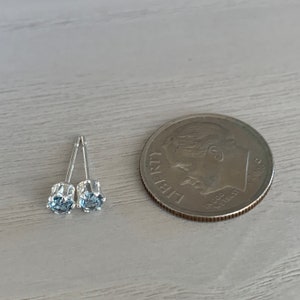 May include: A pair of silver stud earrings with small, light blue gemstones. The earrings are shown next to a US dime for size comparison.