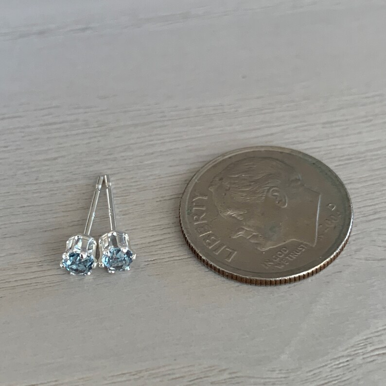 May include: A pair of silver stud earrings with small, light blue gemstones. The earrings are shown next to a US dime for size comparison.
