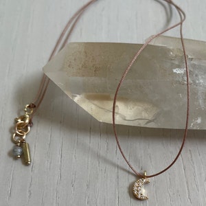 May include: A delicate gold crescent moon pendant necklace with small sparkling stones, hanging from a thin brown cord.
