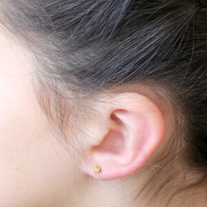May include: A close-up of a person's ear with a small, yellow gemstone stud earring.