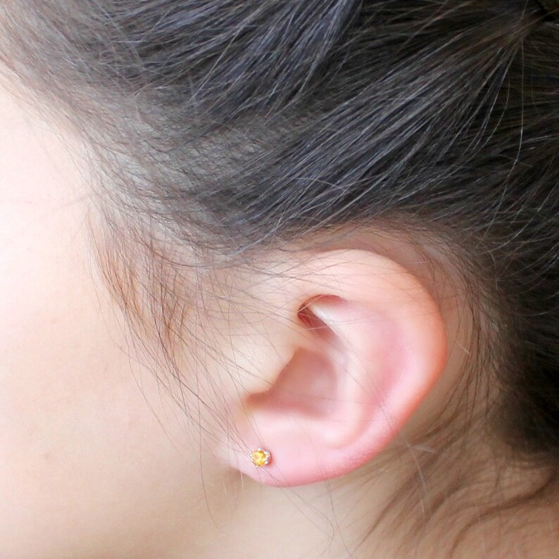 May include: A close-up of a person's ear with a small, yellow gemstone stud earring.