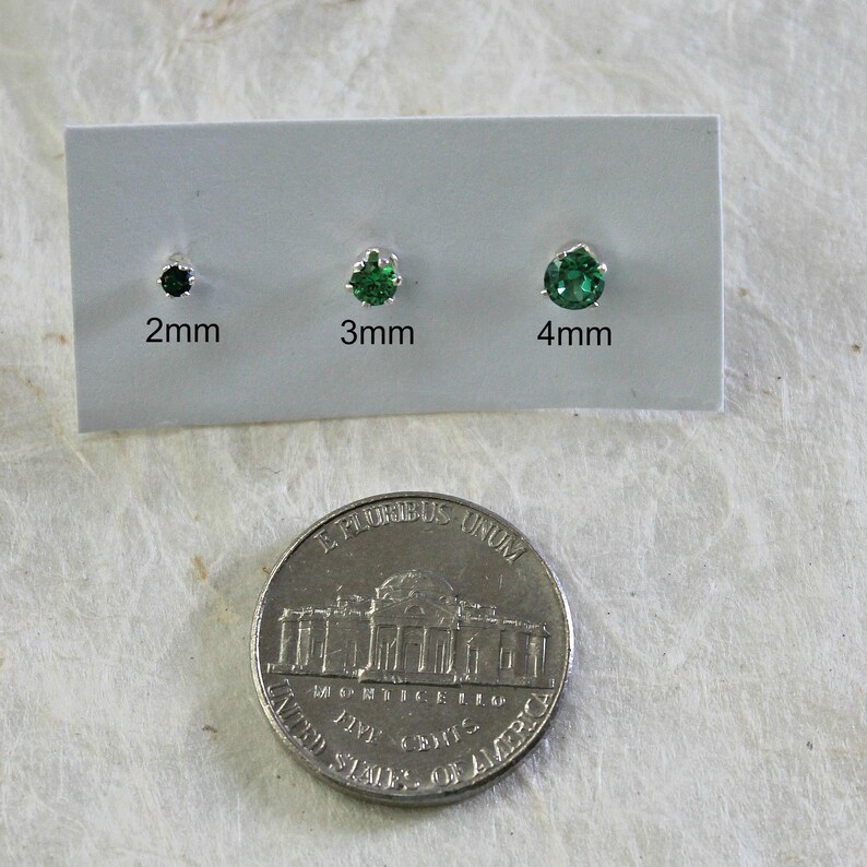 May include: Three green gemstone earrings on a white card with measurements in millimeters: 2mm, 3mm, and 4mm. A US quarter is shown for size comparison.