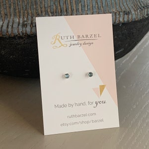 May include: A pair of silver stud earrings with small, round, blue gemstones. The earrings are displayed on a white card with the text "Ruth Barzel Jewelry Design" and "Made by hand, for you." The card also includes the website address "ruthbarzel.com" and the Etsy shop address "etsy.com/shop/barzel".