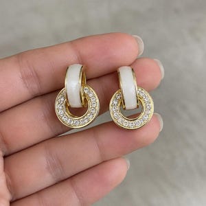 Puede incluir: Un par de pendientes dorados con una pieza superior semicircular blanca y lisa, conectada a un anillo inferior circular incrustado con pequeños cristales claros y brillantes. El diseño combina un acabado metálico pulido con detalles blancos.