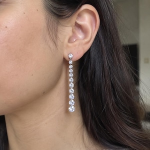 Puede incluir: Un pendiente largo de plata con una línea vertical de diez piedras redondas, transparentes y facetadas que aumentan gradualmente de tamaño de arriba a abajo, creando un diseño lineal y colgante desde el lóbulo de la oreja.