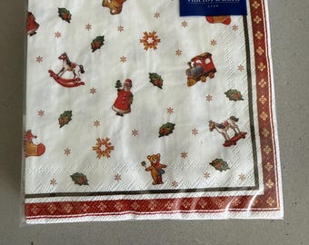 May include: A pack of white paper napkins featuring a Villeroy & Boch 1748 label. The napkins have a festive pattern of teddy bears, rocking horses, trains, Santa Claus, holly leaves, and stars, with a dark red and gold decorative border.