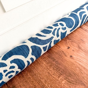 May include: A cylindrical fabric draft stopper featuring a bold, repeating blue and white floral pattern. The textile has a visible woven texture and is positioned horizontally along the base of a white door frame on a wooden floor surface.