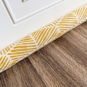 May include: A cylindrical door draft stopper featuring a repeating geometric chevron pattern in mustard yellow and white. The fabric cover has a textured appearance and is positioned horizontally along the base of a white door frame.