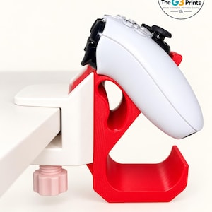 May include: A white and red plastic controller mount clamped to a surface. The mount features a white screw knob and a red hook-shaped arm holding a white gaming controller. A circular logo in the corner reads The G3 Prints Made to Imagine. Printed to Create.