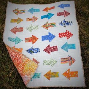 May include: A white quilt with a patchwork design featuring colorful arrows pointing in the same direction. The arrows are made of different fabrics with various patterns, including floral, geometric, and abstract designs.