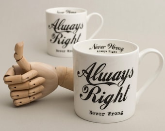 May include: Two white ceramic mugs with black lettering. The mugs say "Always Right Never Wrong". One mug is in focus and the other is out of focus. A wooden hand with a thumbs up is in the foreground.