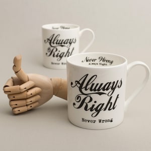 May include: Two white ceramic mugs with black lettering. The mugs say "Always Right Never Wrong". One mug is in focus and the other is out of focus. A wooden hand with a thumbs up is in the foreground.