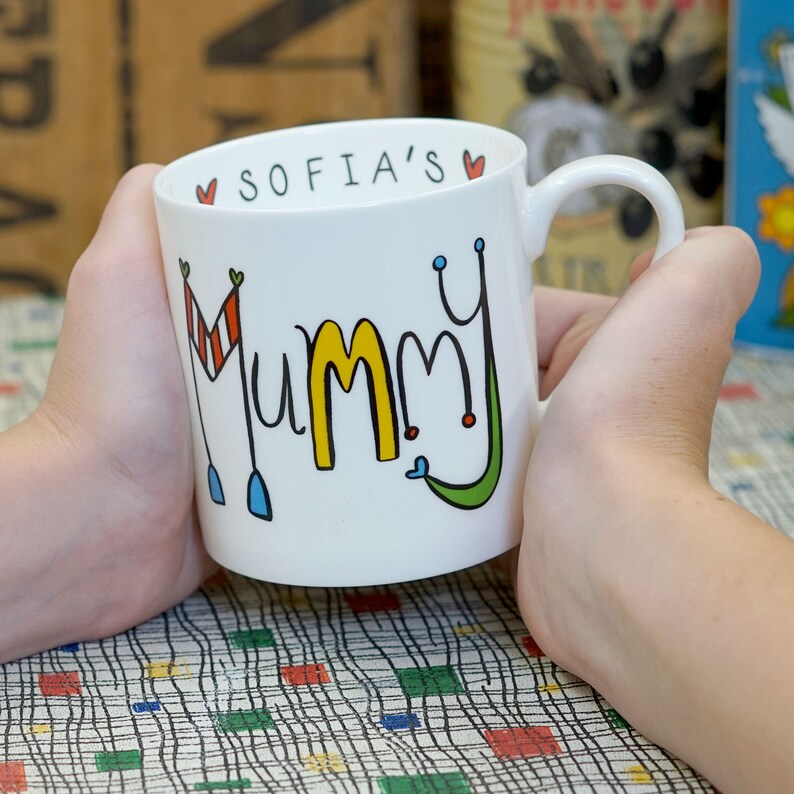 May include: White ceramic mug with a colorful "Mummy" design and the text "Sofia's" on the rim. The mug is being held by a hand.