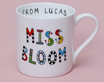 May include: White ceramic mug with a black handle. The mug has the words "From Lucas" printed on the top rim and "Miss Bloom" printed in colorful, stylized letters on the front.