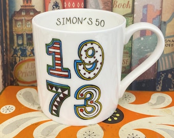 May include: White ceramic mug with a colorful 1973 design and the text "SIMON'S 50" on the front.