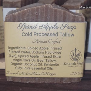 May include: A rectangular bar of brown Spiced Apple Soap wrapped in a tan paper label. The label text reads Spiced Apple Soap, Cold Processed Tallow, Artisan Crafted, and lists ingredients including beef tallow and bentonite clay.