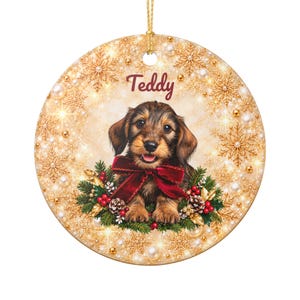 Puede incluir: Un adorno redondo de cerámica con un cachorro de perro salchicha de pelo duro marrón con un lazo rojo. El fondo muestra copos de nieve dorados, perlas blancas y ramas de pino con bayas rojas. El nombre Teddy está impreso en rojo oscuro.