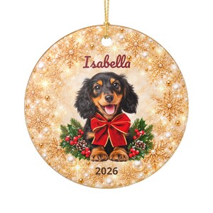 Può includere: Una decorazione natalizia rotonda in ceramica con un cucciolo di bassotto a pelo lungo nero e focato con un fiocco rosso. Lo sfondo mostra fiocchi di neve dorati, perle e agrifoglio. Il testo Isabella e 2026 è stampato sull'ornamento.